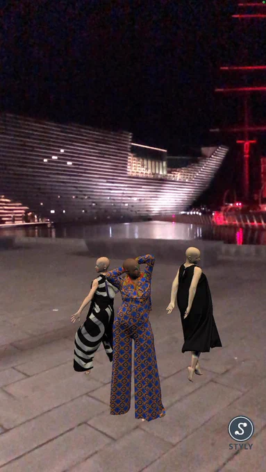 Three avatars - one in a black and white trouser and top suit, one wearing a blue and orange patterned jumpsuit and one wearing a black sleeveless dress - on a pavement outside an in comic museum at night.
