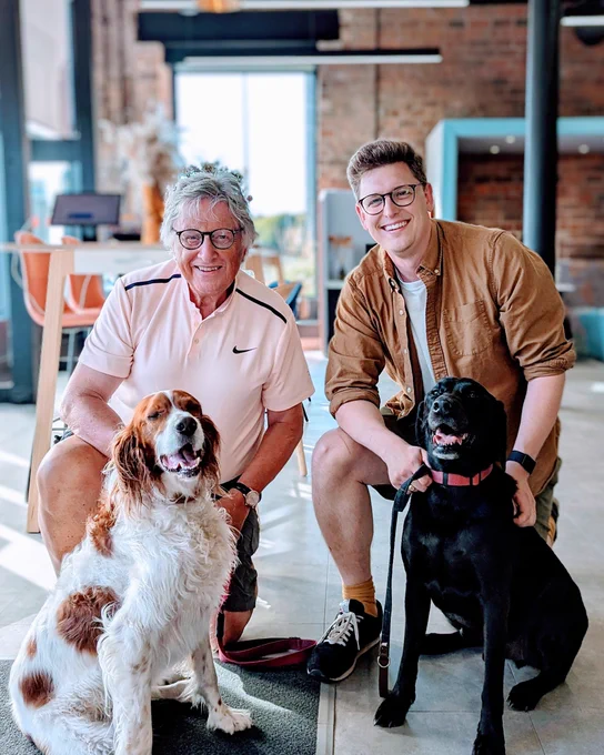 Alistair and Dan both wearing shorts, kneeling behind their dogs Ciara an Irish Red and White Setter and Samwise a Black Labrador. 