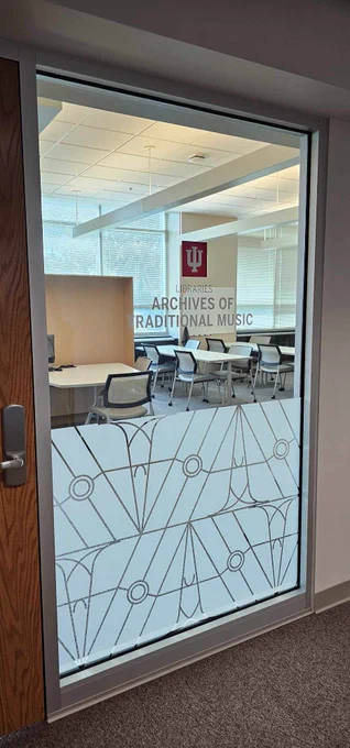 Picture of tall glass window next to ATM door. On window are clings which read "Libraries" and "Archives of Traditional Music" with a graphic frosted design on the bottom half underneath. Through the window there are desks and chairs in the reading room.  