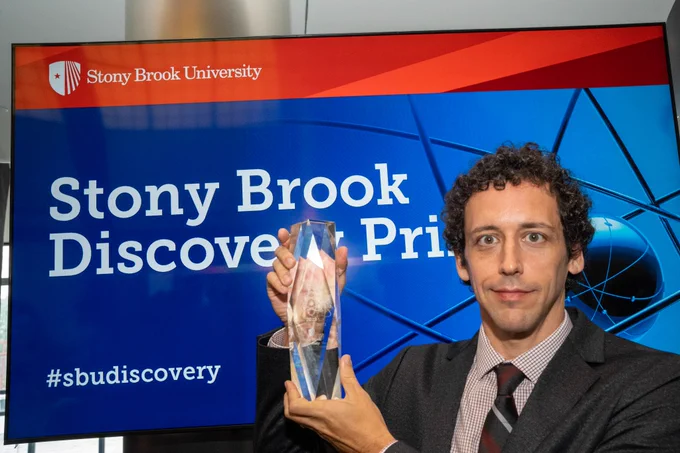 Chris Johnson holding a glass trophy in front of a screen that has a slide with "Stony Brook Discovery Prize" on it.