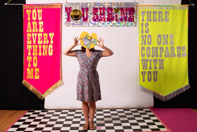 A woman wearing a patterned dress holds a yellow heart in front of her face, featuring an illustration of a rollerskate in lilac, green & orange. She is stood on a black & white checked floor & behind her is a backdrop of a pink & silver sparkly banner featuring the words YOU SHRINE. To the left is a large pink fabric banner that reads YOU ARE EVERYTHING TO ME. On the right is a large yellow banner that reads THERE IS NO ONE COMPARES TO YOU.