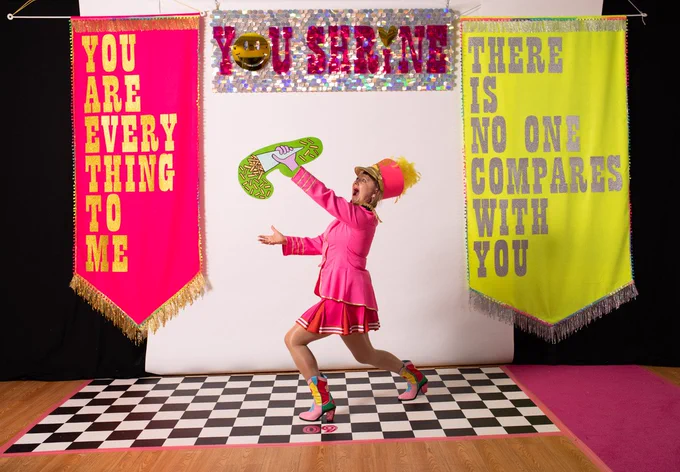 A female wearing a bright pink marching jacket & skirt complete with multi-coloured cowboy boots & pink & yellow feathered marching hat poses for a picture. She holds a cardboard illustrated cut out of a hand holding a cone of chips. She is stood on a black & white checked floor & behind her is a backdrop of a pink & silver sparkly banner featuring the words YOU SHRINE. To the left is a large pink fabric banner that reads YOU ARE EVERYTHING TO ME. On the right is a large yellow banner that reads THERE IS NO ONE COMPARES TO YOU.