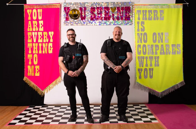 Two male police officers dressed in their black uniforms smile & stand on a black & white checked floor. Behind them is a backdrop of a pink & silver sparkly banner featuring the words YOU SHRINE. To the left is a large pink fabric banner that reads YOU ARE EVERYTHING TO ME. On the right is a large yellow banner that reads THERE IS NO ONE COMPARES TO YOU.