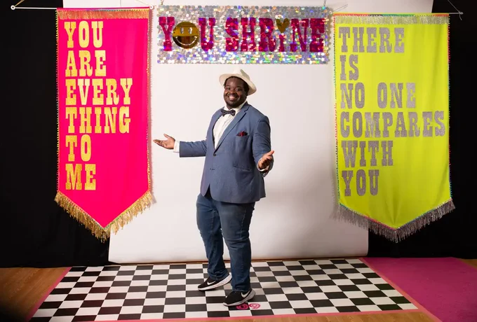A male dressed smartly in a blue suit, bow tie & cream trilby hat poses for a photograph. He is stood on a black & white checked floor & behind him is a backdrop of a pink & silver sparkly banner featuring the words YOU SHRINE. To the left is a large pink fabric banner that reads YOU ARE EVERYTHING TO ME. On the right is a large yellow banner that reads THERE IS NO ONE COMPARES TO YOU.