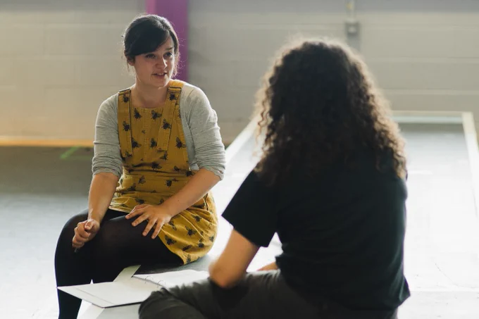 Director Ruby Clarke in rehearsals for We Could All Be Perfect. Photo by Becky Payne. Two women sit on the edge of a raised platform and are in conversation. One wears black and the other wears a yellow pinafore.