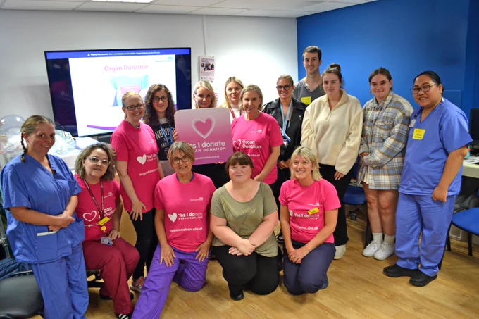 A group of RUH staff members stand together in a seminar room. Some of the staff are wearing blue scrubs and some staff are wearing bright pink 'organ donation' t-shirt. A sign that reads 'Yes I donate' is held.