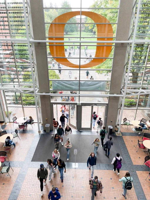 Photo of the atrium of the Lillis Business Complex, taken looking down from a higher floor towards the large glass windows of the building's south entrance, where students enter and exit beneath the large yellow "O" sticker above the doors.