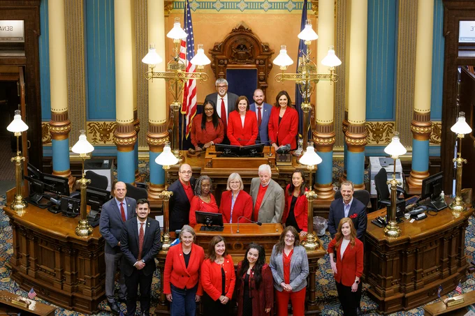 Michigan's Democratic State Senators standing in rows at the beautifully ornate rostrum in the front of the Senate chamber. The Senators are smiling and wearing shirts, button-ups, blazers and ties in the color red to show their solidarity with the UAW members on strike.