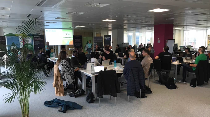 View of an open office space with around 30 people sat at tables listening to a presentation, with plants around the edges