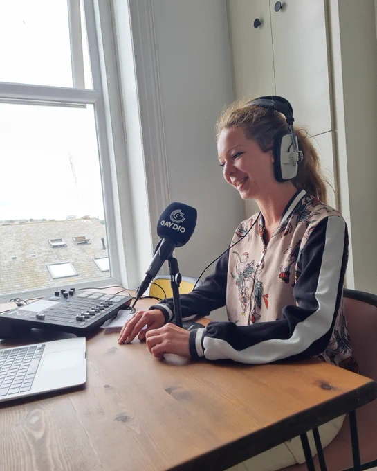 A person sits in front of a radio mic, wearing headphones. There's a laptop and mixer on the desk. 