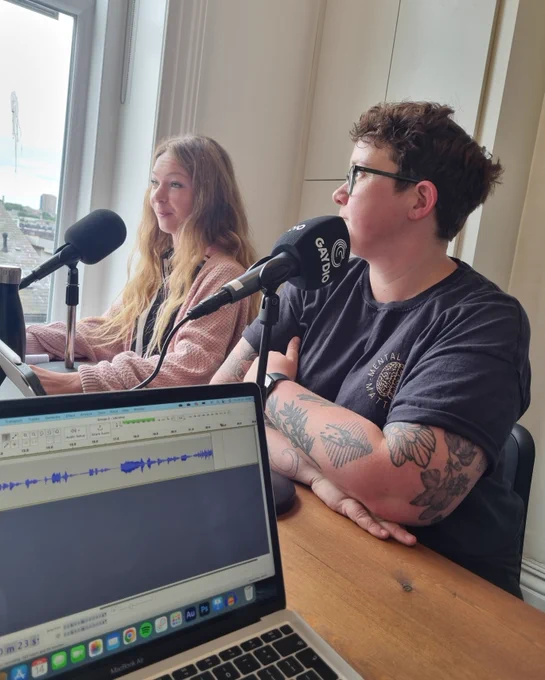 Two people sit in front of microphones. There is a laptop with an audio wave in the foreground.  