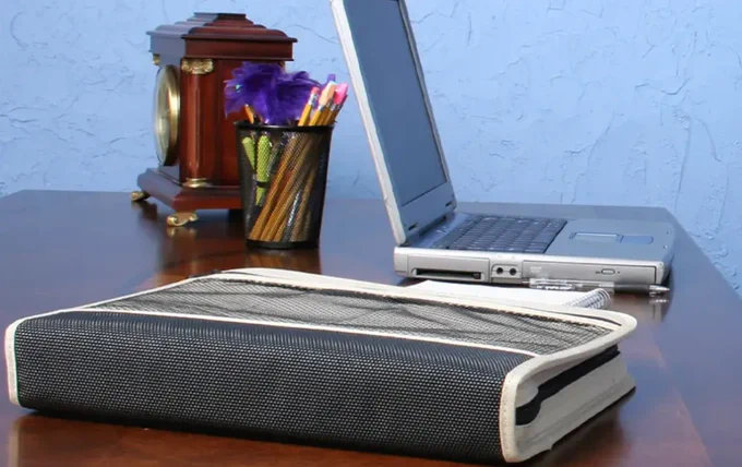 Image of a zipped binder, laptop, clock, and cup of pens, pencils, and highlighters sitting on a desk.