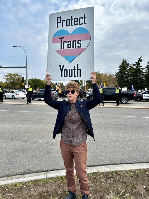Janis holds a sign that reads Protect Trans Youth with a backdrop of protesters