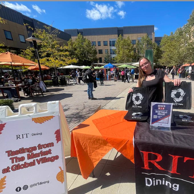 The picturesque Global Village setting with outdoor tables and umbrellas is shown beneath a beautiful blue sky. In the foreground, a sign reads "Tastings from The Market at Global Village" next to a table showing off RIT Dining swag.