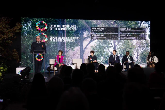 Prime Minister Justin Trudeau sits at an armchair beside four other people on stage. They are looking towards the Fire Chief of West Kelowna, Jason Brolund, who is speaking at a podium in front of a crowd. A projection behind them displays the event title, “FROM THE FRONT LINES OF THE CLIMATE CRISIS.”