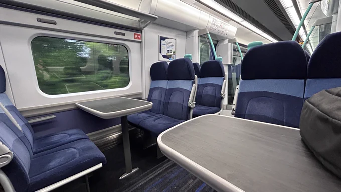 Interior of a train: dark and light blue seats, black and blue striped carpet and black tables in the middle of a four seat formation.