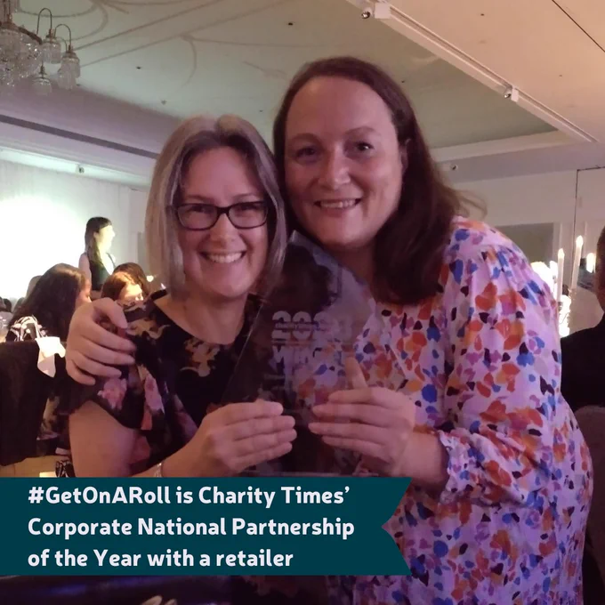 Image of Cara Hoofe, Marks & Spencer employee and bowel cancer patient advocate and Nicolette Barton, Head of Marketing and Communications at Bowel Cancer UK. They're holding the award and smiling at the camera. On top of the image is a ribbon which says: "#GetOnARoll is Charity Times’ Corporate National Partnership of the Year with a retailer".