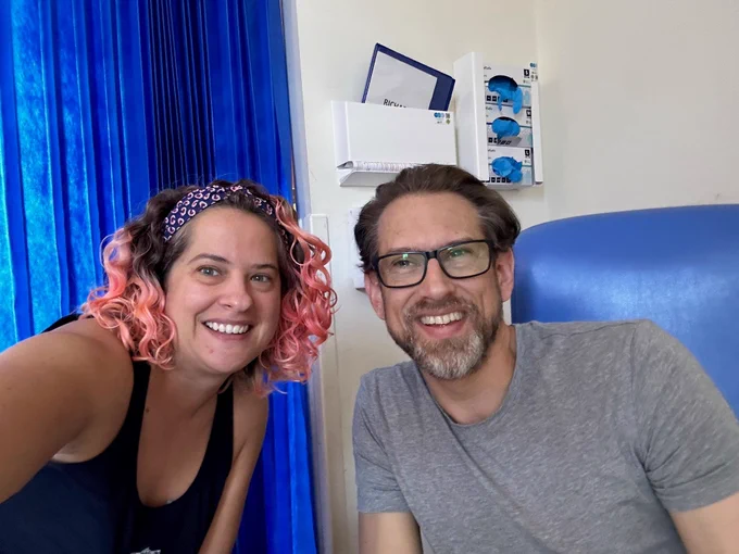 A photo of Imogen Levy on the left and her brother Graeme on the right. Imogen has pink curly shoulder length hair tied in a blue and pink hairband and wears a black sleeveless top. Graeme is sitting in a blue hospital chair and wears black glasses and a grey T-shirt. Behind them is a blue hospital curtain which is drawn. A hospital chart and blue gloves are in white containers on the wall. Both are smiling.