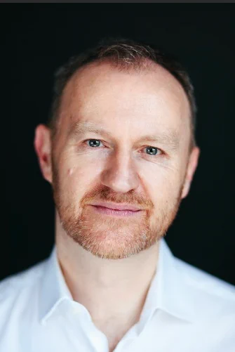 A headshot of Mark Gatiss. He is a 56-year-old white man who has a trimmed ginger beard and mustache. He is wearing a white shirt and looking in the camera.