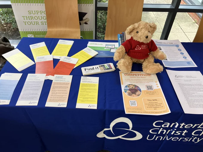 A lovely teddy bear, alongside information leaflets on a blue tablecloth with the Canterbury Christ Church University logo.