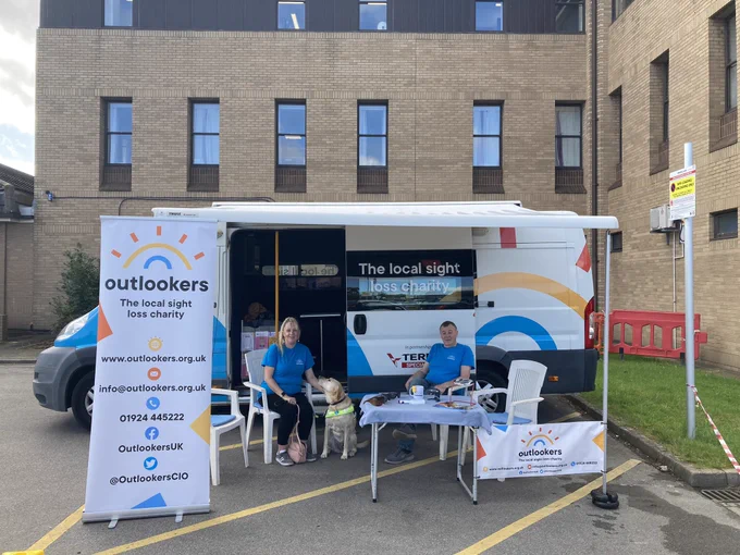 The mobile unit is parked up with its canopy out a gentleman and lady with a guide dog, or seated on chairs next to a table beside the van