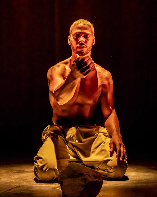 Production photo of Ira Mandela Siobhan, kneeling and looking at the palm of his hand.