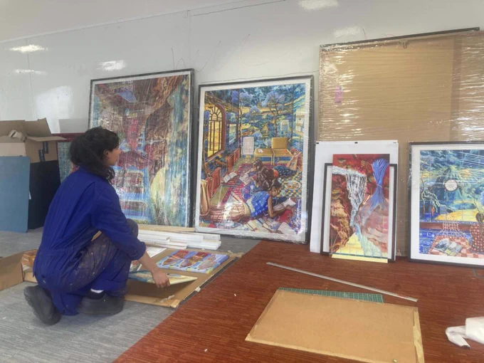 A photograph of someone in blue overalls kneeling down to place multiple large paintings against a wall. 