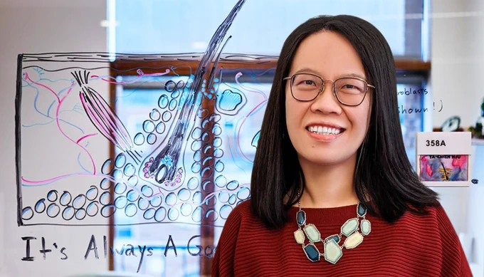 Woman with shoulder length black hair wearing a red sweater stands in front of a glass window displaying a hand-drawn illustration