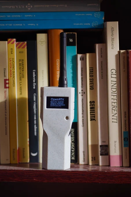 Picture of a LILYGO T-TWR Plus radio standing on a bookshelf showing the OpenRTX about screen with the main authors’ names.
