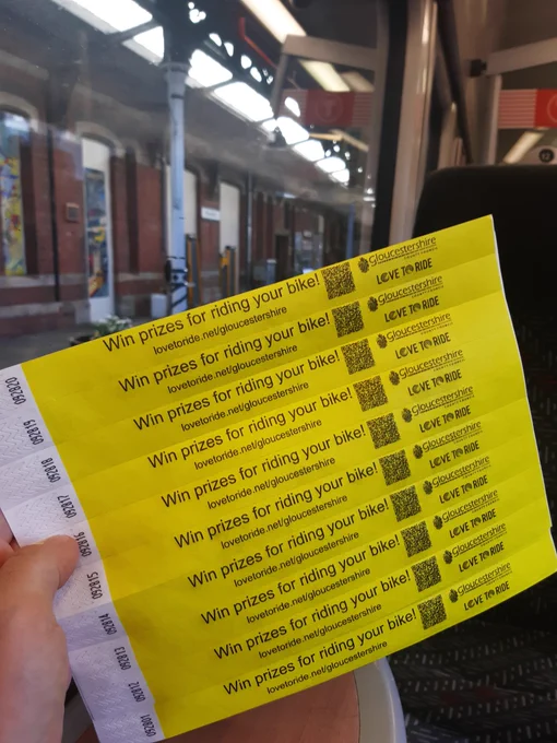 Yellow paper printed bands promoting Love to Ride are held up in front of the train window heading as the train pulls up to Gloucester station.
The bands display the text "Win prizes for riding your bike!"

They also carry the love to Ride and Gloucestershire council logo a QR code and the Love to Ride website. 