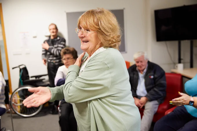 A group of older people are gathered together at a dance workshop. A woman in the foreground crosses one arm across her body and is smiling. A man in the background is doing the same action whilst others, who are seated, watch.