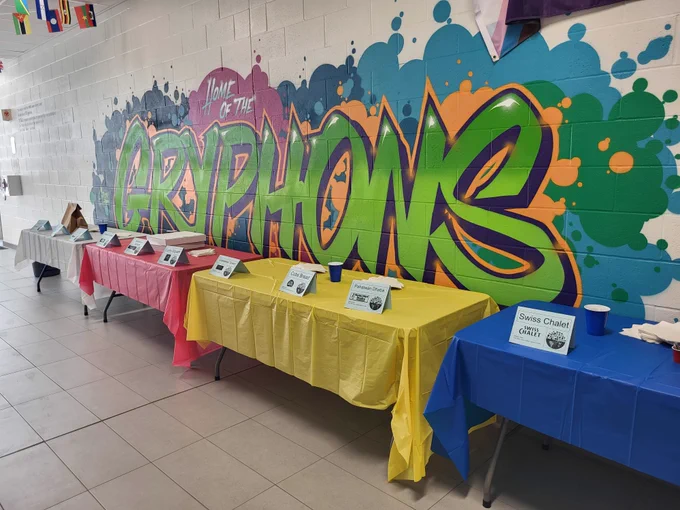 A hallway inside a school. On the wall there is a graffiti-style mural that says "Home of the Gryphons" in neon green letters. In front of the wall are four tables with multicoloured tablecloths that are set up to present a variety of multicultural foods.