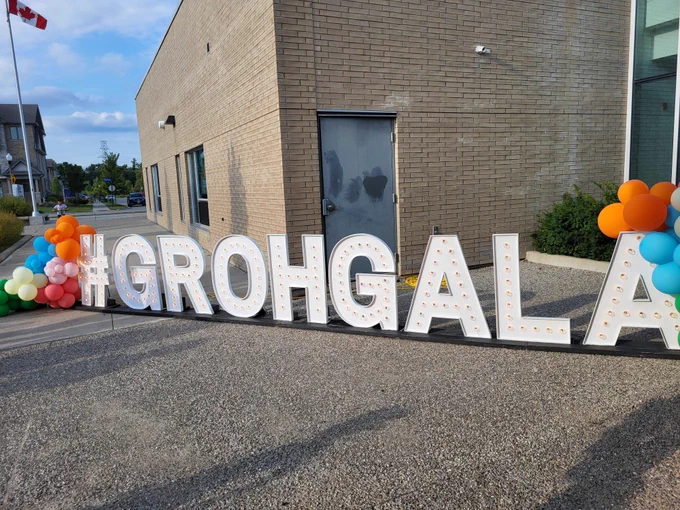 A school with a sign in front of it that says #GrohGala. The sign has orange, blue, and pink balloons on each end of it. The sign's letters are made of lightbulbs.