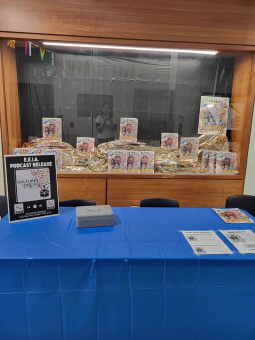 A showcase at a school with approximately one dozen copies of a book called "Deliciously Different." In front of the showcase, a table with a blue table cloth is set up to do book signings. On the table there is a poster advertising the "Cultures Unboxed" podcast.