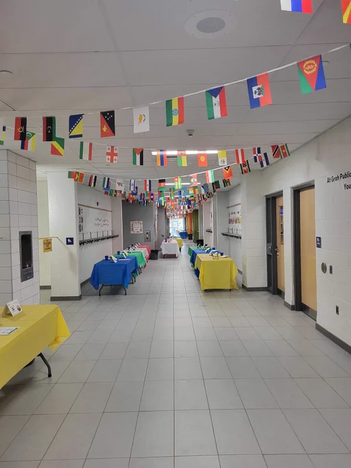 A hallway in a school. On the ceiling is a long garland with world flags hanging down. The hallway has many tables set up with multicoloured table cloths, set up to showcase food from cultures around the world. 