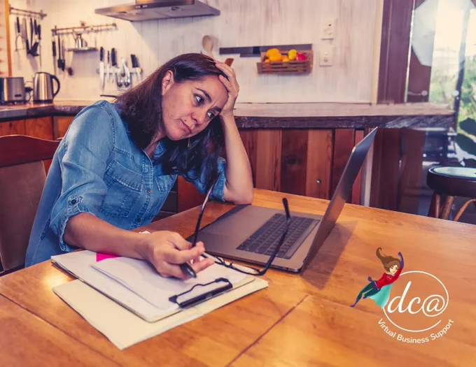 How Work-From-Home Models Have Impacted Our Mental Health. Image of a woman working from home looking stressed at her laptop while sitting at a table in her kitchen. DCA Virtual Business Support.