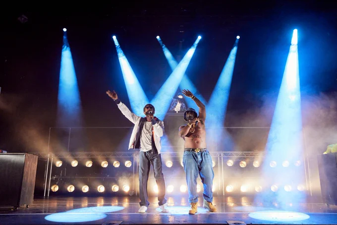 Rhashan Stone and Daniel Ward stand in front of a metal stage structure with lights below and spotlights visible from above. They both have one arm in the air, swaying them to music, and both hold microphones, performing for the audience. Behind them is a masked DJ at a booth above. Rhashan wears jeans and a white jacket with heavy chains. Daniel is shirtless with jeans and a denim bucket hat. 