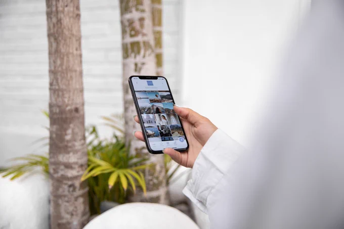 A hand holding an iPhone looking at photos, in a white room with plants in the background.