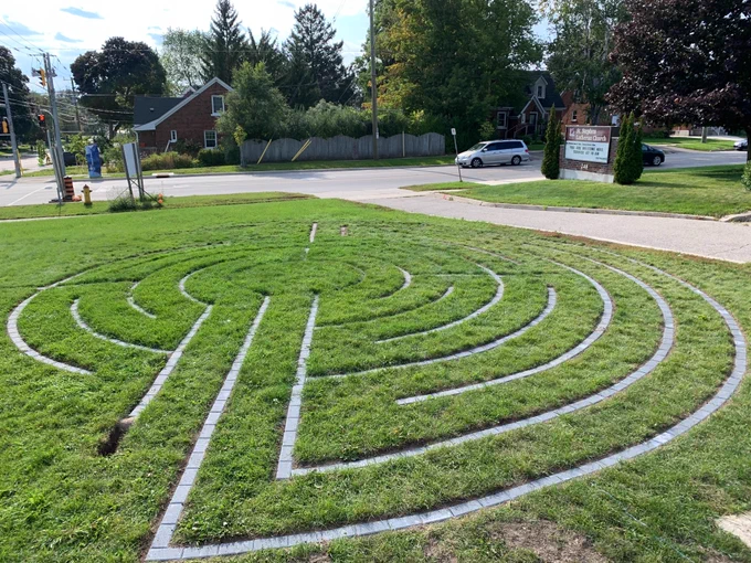A circular labyrinth, made of paving stones set into a lawn, is being built. The area to the right is finished, the left needs more work, and the middle five rings of stone have been completed.