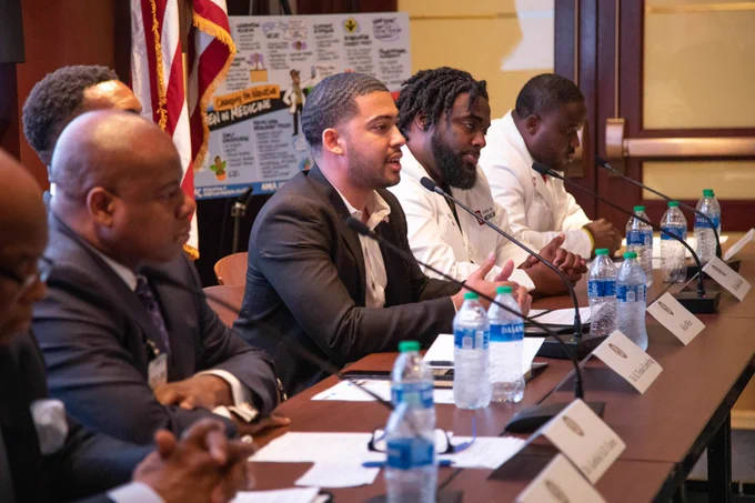 Kylar Wiltz seated in the middle of six other panelists and speaking on the at the April 27th Briefing on Black Men and Boys in Medicine.