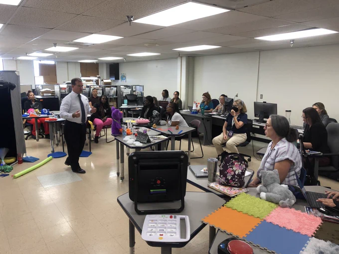 Dr Ray speaking to a group about the importance of Sensory, Literacy & Access.  Room has individuals sitting and all sorts of solutions to support student needs around the room