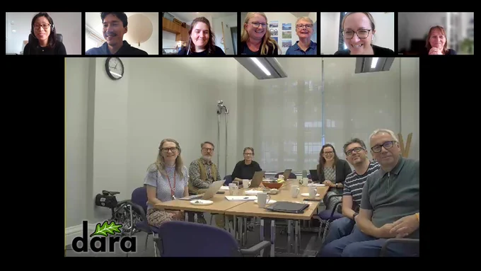 Screenshot of a zoom call. 13 people are shown smiling at the camera from different Zoom windows. In the largest speaker view shot, 6 people are seated around a meeting table. Everyone looks happy. 