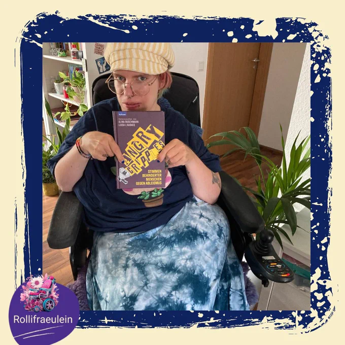 ich sitze in einem Rollstuhl und  halte ein Buch mit dem Titel “Angry Cripples” von Leykamp Velag. Ich trage ein blaues T-Shirt und einen blauen Batikrock. ich sitze in einem Raum mit einem Holzboden und vielen Pflanzen im Hintergrund.