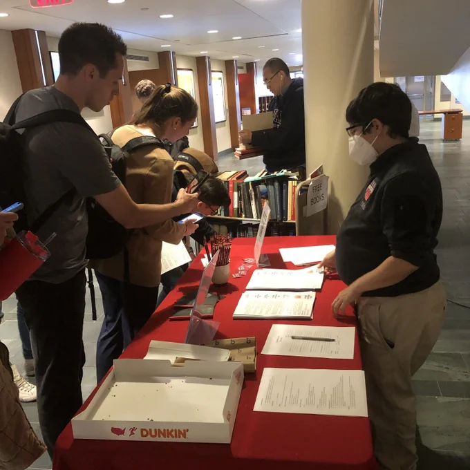 An HLS Library staff member stands behind a table covered with a red tablecloth. An array of library information and giveaways are on the table, along with a (temporarily empty) box of donuts. Students stand in front of the table looking at the information. In the background is a cart of free books.