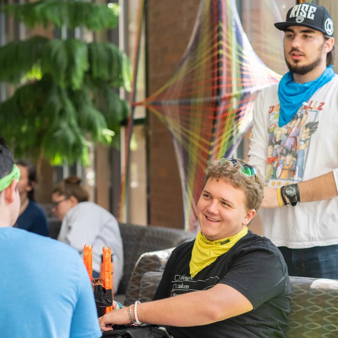 Three young men are gathered, talking. The one of the left is sitting with his back facing the camera and wears a green bandana around his head. The one in the middle is sat smiling, wearing a yellow bandana around his neck and is resting his foam blaster beside himself. One man stands, wearing a blue bandana around his neck.