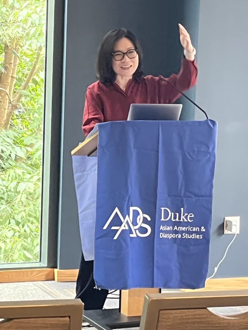 Esther Kim Lee, smiling, gestures toward a colleague from behind a lectern covered in a blue cloth with the text "Duke Asian American & Diaspora Studies"