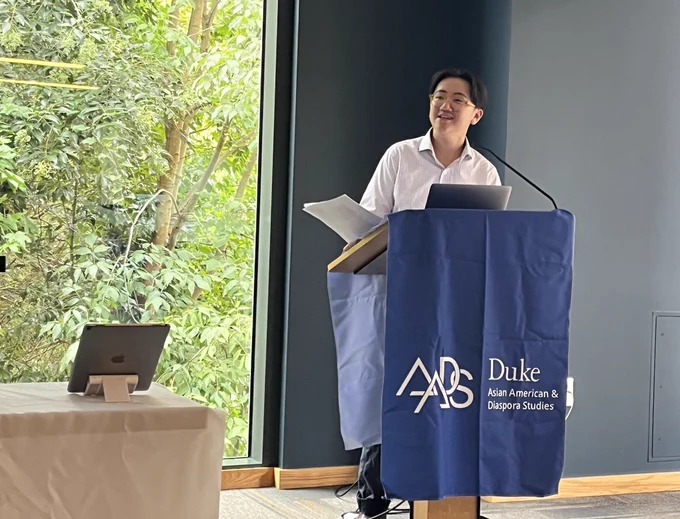 Emily Lim Rogers, holding papers, speaks from behind a lectern covered in a blue cloth with the text "Duke Asian American & Diaspora Studies"