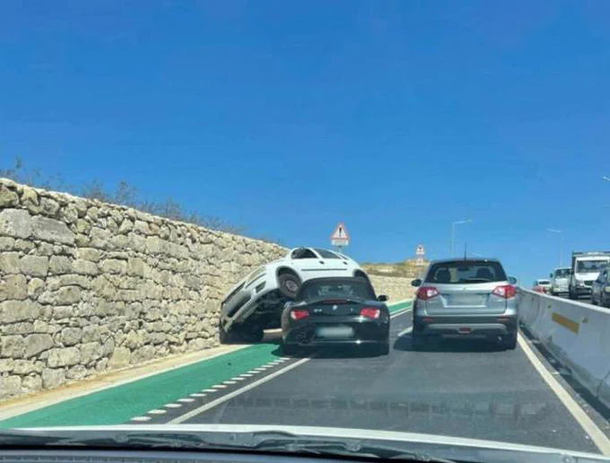 Photograph of a car collision with a white car on top of a black car, both of which are in a green cycling lane that offers cyclcsts no protection from cars.