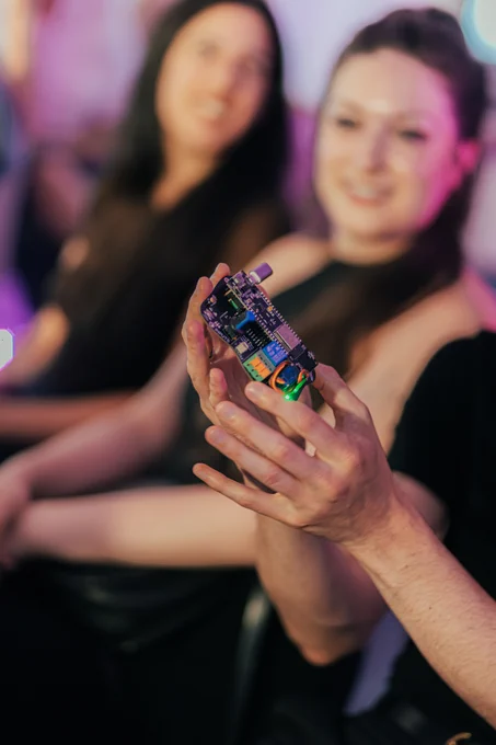 Hands hold a custom microcontroller. Most of the parts are black and silver except the servo and relay are bright blue, green and orange.
