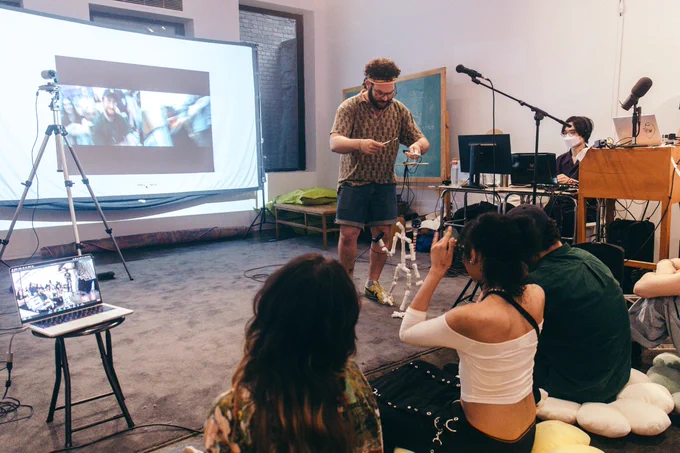 A person doing pipe puppetry in front of audience. Behind them is a projector screen showing the live webcam from the puppet. 